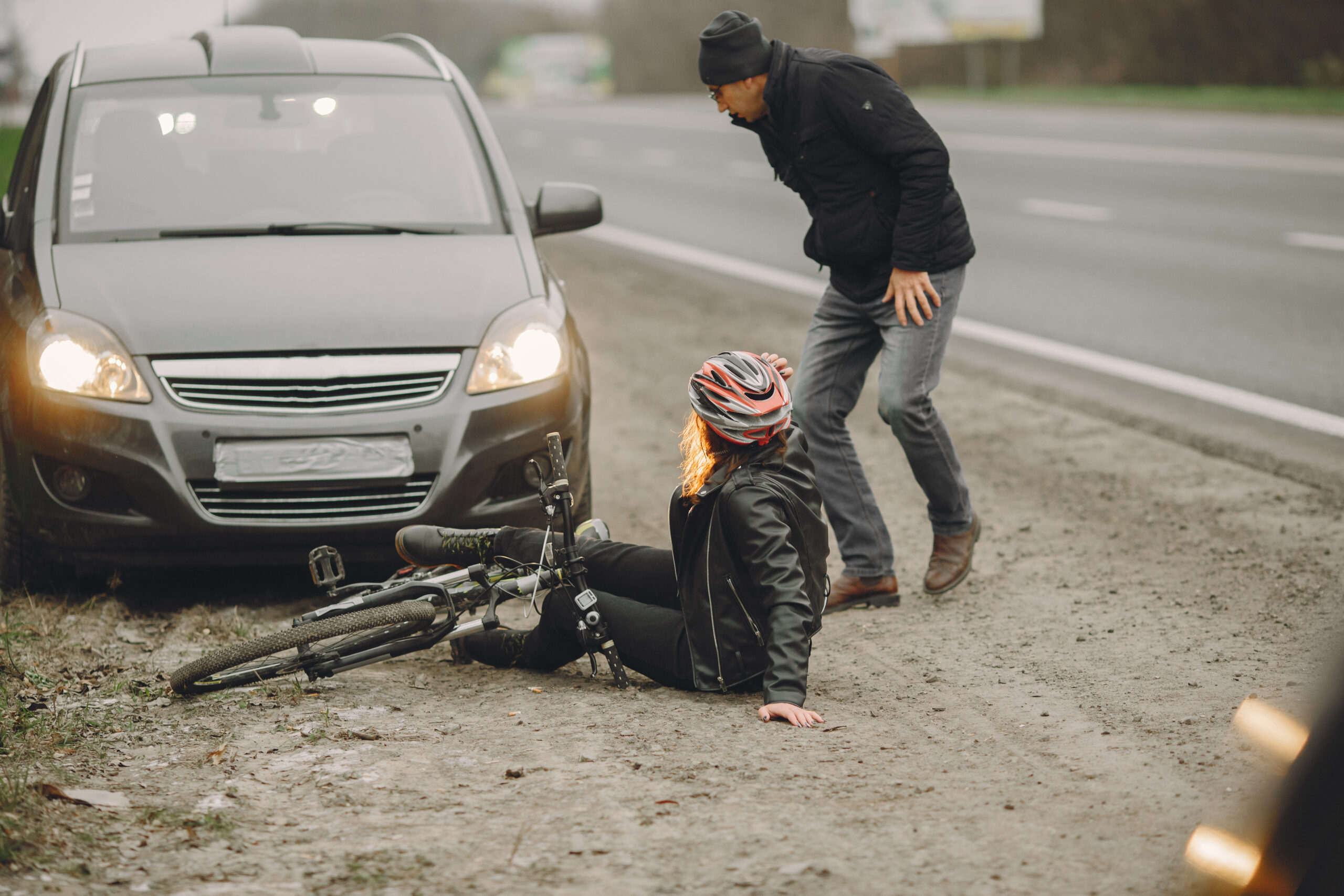 Ciclista caída no chão ao lado de uma bicicleta após possível colisão com um carro parado com os faróis acesos; uma pessoa se inclina para verificar a situação.