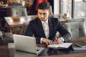 Homem de terno sentado à mesa em um ambiente corporativo, trabalhando com notebook e documentos enquanto faz anotações com uma caneta.