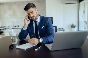 Homem de terno azul fala ao celular enquanto faz anotações em um caderno ao lado de um notebook, em um ambiente de escritório moderno.