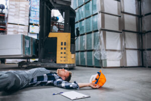 Trabalhador caído no chão de um armazém ao lado de uma empilhadeira, com o capacete de segurança no chão após um aparente acidente de trabalho.